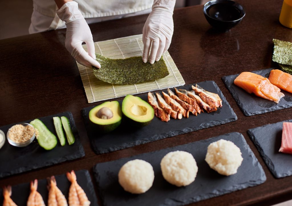 Close-up view of process of preparing rolling sushi. Chef's hands are holding sheet of nori. Ingredients: cucumber, salmon, rice, avocado on black stone plates